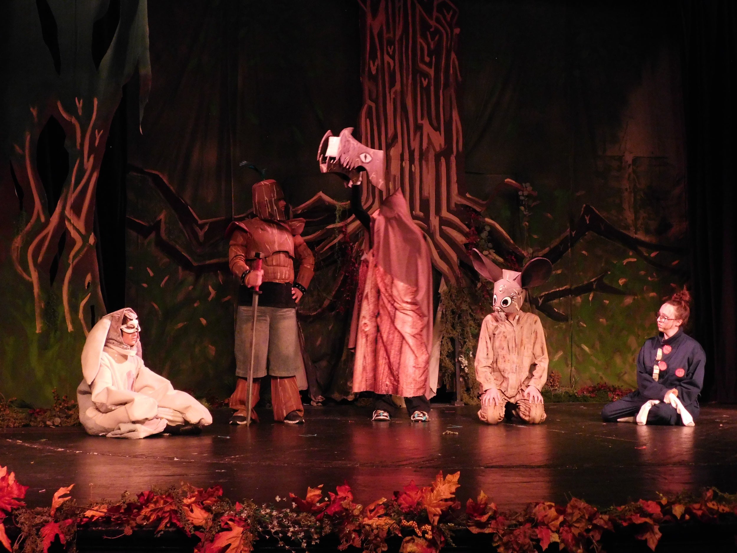 Stage performance with costumed characters in front of a dark, tree-like backdrop